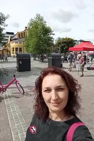 The video shows a woman with curly brown hair standing on a paved sidewalk. She is wearing a dark shirt with a crest on the left chest and a pink backpack strap is visible over her right shoulder. She is looking at the camera and smiling. In the background, there is a street with buildings on either side. Some people are walking on the sidewalk and in the street, and there are bicycles and trash cans visible. The buildings appear to be shops or businesses. The sky is cloudy.