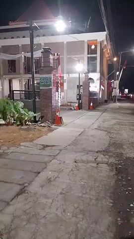 The video shows a nighttime street view with a building on the left and a street on the right. The building has red and white decorations, possibly flags, and lights are on. There is a sign that says "PARKIR MOBIL," which translates to "car parking." The street appears to be paved, with some cracks and debris. Several cars and motorcycles are visible on the street in the distance, with their headlights on. The overall impression is of a street scene at night, possibly in a commercial area.