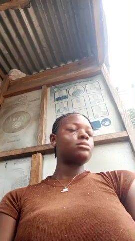 A young woman with dark skin and braided hair stands in an enclosed space, possibly a bus stop. She wears a brown, short-sleeved shirt with small embellishments and a silver necklace. Behind her is a framed informational board with several small portraits and text. The space has a corrugated metal roof supported by wooden beams. The woman looks off to the side, then towards the camera, and smiles slightly. The lighting appears to be bright and somewhat diffused.