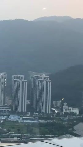 The camera looks out over a landscape with tall apartment buildings in the foreground, a line of green hills in the middle distance, and a hazy sky with a sunset. The sun is a bright orange disk peeking through the clouds. The apartment buildings are tall and gray. The hills are covered in green trees. The sky is a pale gray.