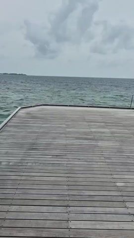 The video shows a wooden pier extending out into the ocean. The pier is made of planks of grey wood and the ocean is teal. There is a building on the pier with a brown roof and brown walls. The building has a white door and the number 35 on it. The sky is overcast and the horizon is visible in the distance. The camera pans from the pier to the building and back to the pier. The camera remains stable throughout the recording.