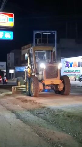 This media file captures a yellow grader operating on a dirt road at night. The grader is the primary object, positioned centrally in the frame and actively engaged in grading the road surface. The scene is illuminated by the grader's headlights and other ambient light sources from nearby buildings and signs.

To the left, a brightly lit sign for "Indomaret" is visible, indicating a convenience store. Below it, another sign reads "PAKET" with a blue background, likely advertising postal or delivery services. Further left, a sign with the word "mangga" is partially visible.

On the right side of the frame, a large, colorful sign with the word "STAR" and a star graphic is prominent. Text beneath it reads "KEPUASAN ANDA KEBANGGAAN KAMI". Below this, a website address is visible: "starkids.go.id".

The setting appears to be a road in Blangpeuria, Indonesia, given the provided location context. The time of day is clearly night, suggested by the darkness and the artificial lighting. The weather appears clear, as there are no visible indications of rain or adverse conditions. There are no people clearly visible interacting with the grader, though a person might be inside the cabin. The overall mood is one of ongoing work and infrastructure development in a semi-urban or commercial area. The grader's bright lights cut through the darkness, highlighting the dirt and gravel of the road it is smoothing.