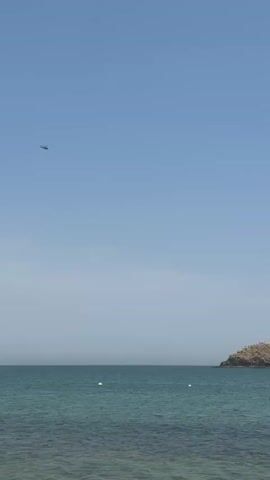 A helicopter flies across a clear blue sky above a serene body of water, likely the sea or ocean. In the distance to the right, a small, rocky island is visible, appearing to be covered in birds or vegetation. Two white buoys float on the water's surface in the foreground, suggesting a designated area or potential markers. The water itself is a clear turquoise color, indicating shallow or calm conditions. The overall scene suggests a bright, sunny day at a coastal location, possibly Al Fiqait, United Arab Emirates, given the provided context. No other specific activity or text is visible.
