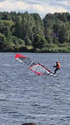 A person is windsurfing on a body of water. The person is wearing an orange life vest and black pants. They are standing on a white board and holding onto a sail. The sail is red and black. The water is choppy. In the background, there is a forest and a cloudy sky.