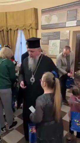 In a well-lit indoor setting with a checkered floor, a bearded man in a black cleric robe and hat interacts with a young girl with Down syndrome. The girl wears a dark, glittery dress and pink glasses, and holds a small religious icon and a blue gift bag with stars. The cleric is handing out items, possibly gifts. Several other people, including women and children, are present, some holding similar gift bags. One man in a blazer stands in the background. The walls are decorated with framed displays. The cleric poses for a photo with the girl, both smiling at the camera.