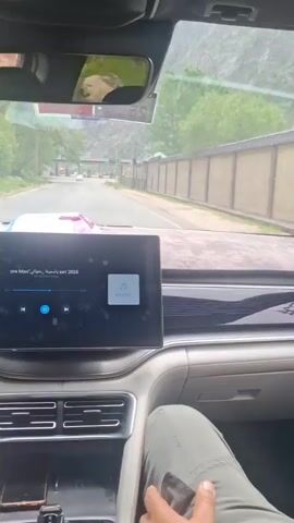The camera is positioned inside a car, providing a view through the windshield toward what appears to be a border crossing or toll booth. In the foreground is the dashboard of the car, which features a large touch-screen display showing music controls. Through the windshield, multiple lanes with traffic lights are visible, some displaying green and others red. There are also signs posted with traffic information. Outside, the landscape appears to include mountains and a wall running along the side of the road. The day seems overcast.