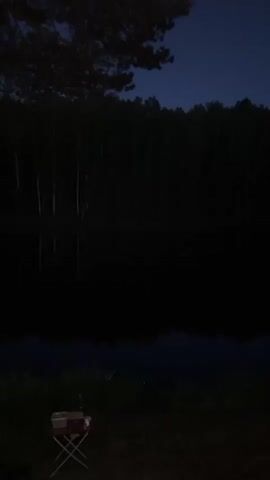 A handheld video captures a dark, evening or night scene of a calm body of water with a thick forest on the opposite bank. The water reflects the dark sky and the trees. The camera slowly pans to the right, revealing more of the shoreline. The sky above the forest is a deep blue, suggesting twilight or early night. In the foreground, on the grassy bank, there's a small, foldable camping chair and some bags or items on the ground. Two tall, slender tree trunks with sparse foliage are visible on the right side of the frame as the camera continues to pan. The overall ambiance is tranquil and dark.
