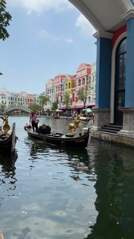 The video shows a picturesque scene featuring a gondola on a canal, reminiscent of Venice, but set against a backdrop of brightly colored buildings.

In the foreground, a black gondola is positioned on the calm, reflective water. It has a distinctive golden statue at its front, depicting a centaur-like figure with a bow drawn, aiming forward. On the side of the gondola, text is visible, including "E.0.4" and "KG-00007". A gondolier, dressed in a striped red and white shirt and a hat, stands towards the back of the boat, holding a long oar. He appears to be maneuvering the gondola, which is either stationary or moving very slowly alongside a building. Another gondola is partially visible on the left side of the frame.

The canal itself is surrounded by vibrant, multi-story buildings that line its banks. These structures display a lively palette of colors, including reds, yellows, oranges, blues, and greens, contributing to a cheerful atmosphere. An arched bridge is visible in the distance, spanning the canal, with more colorful buildings beyond it.

The sky is bright and clear, suggesting a sunny day. The reflections of the buildings and the gondola are clearly visible on the water's surface. The overall setting evokes a sense of leisure and travel, characteristic of a tourist destination designed to resemble a European city.