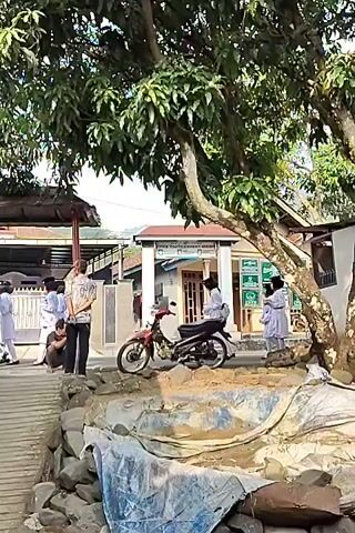 The video shows a group of people standing near a building and a tree. A red and black motorcycle is parked in front of the building. Several people are wearing white uniforms. One person is crouching down. A ramp is visible in the foreground. The ground appears to be uneven and covered with rocks and some blue material. The scene looks like it could be an outdoor area of a school or religious institution.
