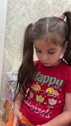 A young girl with long, dark brown hair styled in two pigtails is looking down at a phone she is holding. She is wearing a red t-shirt with green text that says "Happy" and a graphic of bees and flowers. She is also wearing orange shorts. The background appears to be a room with patterned wallpaper and a bed with a duvet and pillows.