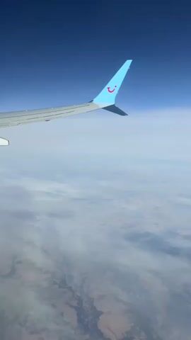 This is a first-person view from an airplane window, looking out at the sky and the landscape below. The right wing of the airplane is prominently featured in the foreground, with a light blue tip displaying a red smiley face logo. The wing extends to the left side of the frame. Below the wing, a vast expanse of clouds stretches across the horizon, obscuring most of the ground. The ground visible through the clouds appears to be a mix of tan and dark brown, with some linear features that could be roads or rivers. The sky above is a deep, clear blue. The video appears to be a continuous shot with no significant changes in the scene.