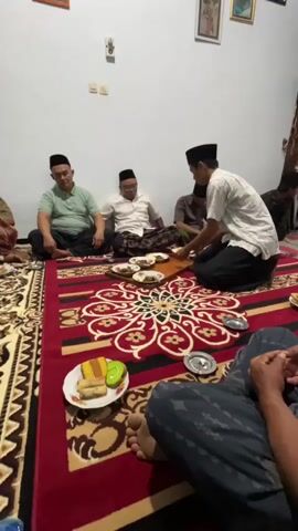 Four men are sitting on a red rug with a circular floral pattern. Three men are seated on the floor with their legs crossed, facing a tray of food in the center. One man, wearing a white shirt and a black cap, is kneeling and serving food from a plate to a smaller plate on the tray. Another man, wearing a light green shirt and black pants, is seated to the left. A third man, wearing a white shirt and black pants with a black cap, is seated in the middle. The fourth man is seated behind the serving man. There are plates of food, including what appears to be cake and spring rolls, on the rug. The room appears to be a home, with a white wall and framed pictures on the wall.