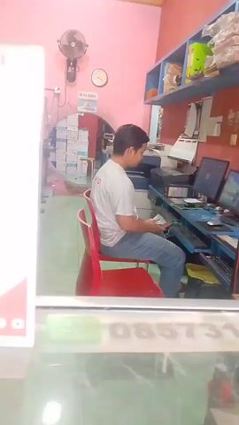 The video shows the interior of what appears to be a small business, possibly a printing or copy shop. In the foreground, there is a glass partition with some signage, including a QR code and what looks like a phone number. Through the glass, a man in a white t-shirt and jeans is seated at a desk with computers and a printer. He is looking down at something in his hands. In the background, shelves are filled with various items, including boxes and bottles. The walls are painted pink and green. Later in the video, another man, wearing a green t-shirt, appears and is holding some papers. He is standing near a large copier machine. The lighting is somewhat dim and the video quality is low.