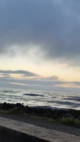 The video captures a static wide shot of an overcast coastal landscape. A grey, cloudy sky dominates the upper portion of the frame, transitioning to lighter, warmer tones of yellow and soft orange near the horizon, suggesting dawn or dusk. Below the horizon, the ocean displays gentle to moderate waves, with some whitecaps visible. In the immediate foreground, a weathered, light grey concrete barrier or wall runs horizontally across the bottom of the frame. Beyond this barrier, a dark, rocky shoreline with sparse green vegetation separates the water from the concrete. The only movement observed is the gentle motion of the ocean waves. The ambient sound consists of ocean waves.