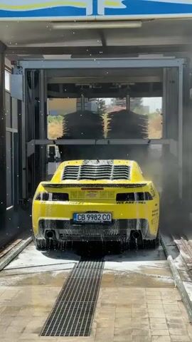 A bright yellow Chevrolet Camaro is captured from the rear in an automatic car wash bay. The car is positioned on a metal grate for drainage, with water and foam being sprayed from overhead nozzles and rotating brushes. The Camaro features black accents, including rear window louvers, a spoiler, and bumper trim with dual exhaust pipes. Its license plate reads "CB 9982 CO". Decals visible on the rear bumper include "CRAZY" on the left and "WE ARE FREE" on the right. The car wash machinery is actively cleaning the vehicle, with water and suds covering its surface. Through the open front of the car wash, parts of buildings and trees are visible in the background under what appears to be a sunny sky. The sounds of the car wash machinery and spraying water are audible.
