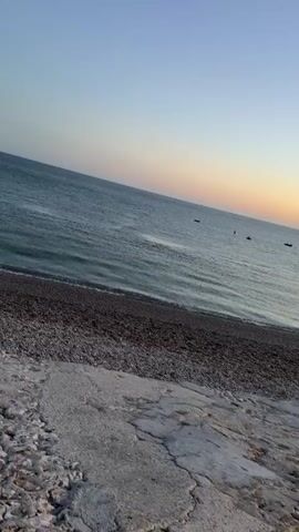 This is a video of a beach at sunset. The sky is clear and a gradient of blue to orange. The ocean is calm with small waves. There are a few small boats in the distance. The beach is made of pebbles and there is a concrete structure in the foreground.