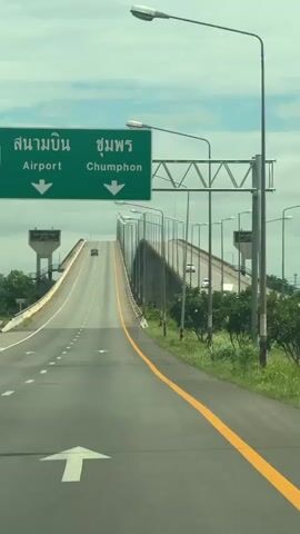 The video shows a two-lane road leading up to a bridge. A green road sign indicates "Airport" and "Chumphon" with arrows pointing downwards. The road is paved and has yellow and white lane markings. Streetlights line the right side of the road. There are a few cars on the road and bridge in the distance. The sky is cloudy.