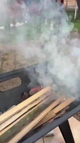 A charcoal grill contains red-hot coals and long wooden planks, emitting thick white smoke. The grill is black metal and positioned outdoors on a tiled surface, with a muddy yard and sparse vegetation visible in the background.