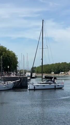 The video opens with a view of a harbor on a bright, sunny day. Several sailboats are docked. In the foreground, a white sailboat with a black canopy is the most prominent. A person, wearing a light-colored shirt, is standing on the deck of this sailboat, appearing to be attending to something. Behind it, another white sailboat is docked. Further back, a large, older-looking sailboat with two masts dominates the scene, moored to the dock. People are visible on the dock near this sailboat, as well as a building with a sign that reads "BAR DU MER". The background features a line of green trees and a building with light blue siding and white trim. The water is calm and reflects the blue sky and white clouds. The camera pans slightly to the right, revealing more of the docked sailboats and the waterfront.