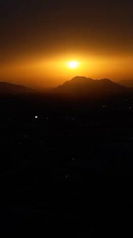 The video shows a sunset over a mountain range. The sun is a bright orange orb at the top of the frame, partially obscured by a large mountain silhouette. The sky is a gradient of orange and yellow, fading to a darker hue at the top. Below the mountains, a dark landscape is visible, with a few scattered lights that suggest a town or settlement. The overall impression is one of a vast and serene natural landscape.
