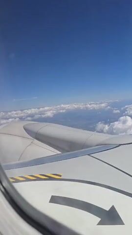 This video shows an aerial view from an airplane window. The wing of the plane is visible in the foreground, and beyond it, there are clouds and land below. The sky is clear and blue. The land appears to be a mix of rural and possibly some small towns or settlements. The clouds are fluffy and white, scattered across the sky.