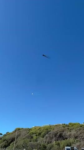 The video captures a clear blue sky with two kites flying. The upper kite is dark and appears to be a stunt kite with multiple tails. The lower kite is smaller and white. Below the kites, a green, tree-covered hill slopes upwards, and a few white buildings are visible near the crest. The bottom of the frame shows a paved area, likely a parking lot, with the tops of several vehicles, including a white van. A power pole with wires is also present on the left side of the frame. The overall scene suggests a sunny day at an outdoor recreational area, possibly a park or beach overlooking a hillside.