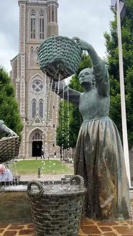 The video features an outdoor fountain with two bronze statues of women. The statue on the right holds a basket above her head, from which water pours into a lower, larger basket on the ground. The statue on the left holds a basket and stands near a rectangular stone basin where water also flows. A man in a light blue shirt is partially visible, leaning over the basin near the left statue. In the background, a large, grey stone church with a tall spire and arched windows stands prominent. Green trees frame the church. Two flagpoles display flags: a French tricolor flag on the left and a black and white striped flag on the right. The ground around the fountain is paved with reddish-brown bricks. The sky is overcast.
