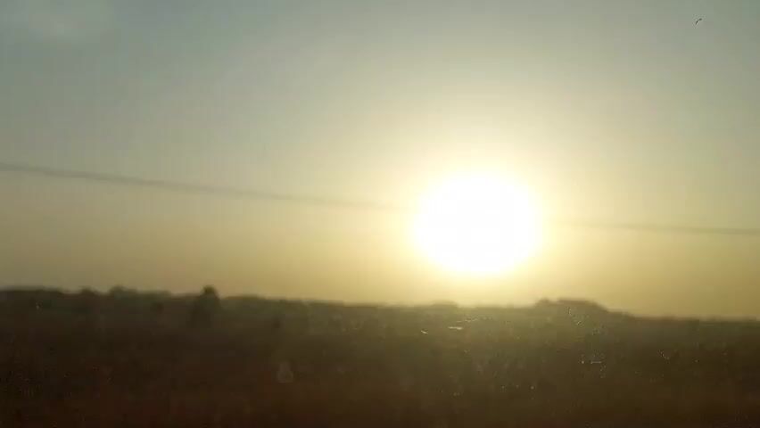 The video shows a hazy sunset with a foreground of blurred landscape and telephone poles. The sun is a bright, round orb in the center of the frame, slightly obscured by the atmospheric haze. A single power line stretches horizontally across the upper part of the view, and a silhouette of land is seen at the bottom. The scene has a muted color palette of yellow and tan.