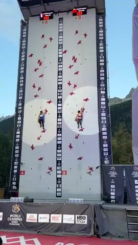 Two competitive rock climbers ascend a vertical climbing wall. The wall is white with numerous red climbing holds. Two digital timers are displayed at the top of the wall, showing 00:00 to 00:05 as the video progresses. Both climbers are wearing harnesses and athletic clothing. The climber on the left is wearing a dark blue top and black pants. The climber on the right is wearing a red top and black pants. Both climbers are actively engaged in the sport, moving upwards on the wall. The environment appears to be an outdoor competition venue, with spectators visible in the background, and banners indicating "WORLD CLIMBING SERIES CHAMPIONSHIPS". Mountains are visible in the distance behind the structure.