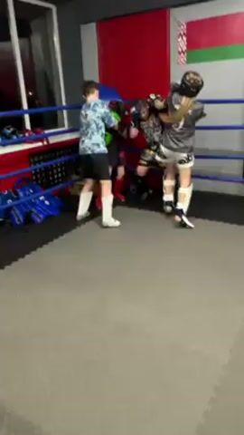 The video takes place in a boxing or martial arts gym, inside a red, white, and blue boxing ring.  Four children or young teenagers wearing protective gear are practicing fighting. Two are actively engaged in sparring in the center of the ring, while the other two appear to be coaches or sparring partners observing from the sidelines.  The environment suggests a dedicated space for combat sports, with gym equipment visible in the background. The room is lit with overhead lights, and the floor is covered with gray interlocking mats.  There is a red, white, and green flag on one wall.