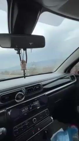 The video is taken from the inside of a car driving on a road. The interior of the car is visible, including the dashboard, steering wheel, and rearview mirror. Outside the car, there is a view of a landscape with green fields, red soil, and hills in the distance. The sky is cloudy and overcast. The camera pans slightly to the right as the car moves along the road. There is music playing in the car.