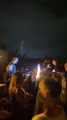 A nighttime outdoor gathering is underway, illuminated by artificial lights and a bright, vertical flame in the center of the frame. A crowd of people is visible, some dancing and socializing. In the foreground, a woman with dark hair is facing away from the camera, holding a drink. To her left, another woman with long, wavy brown hair is partially visible. Behind them, a man with dark hair and braids is looking to his left, holding a yellow cup. Further back, a man wearing a white shirt and a red hat is seen. The background is dark, with faint outlines of trees and structures. The overall atmosphere appears to be festive.