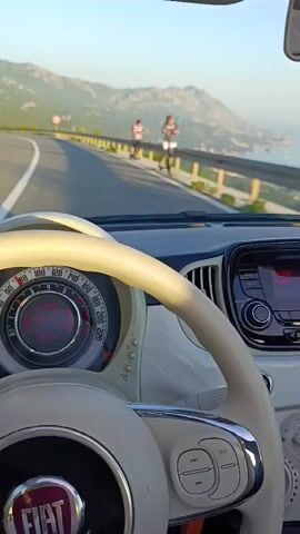 The video shows a first-person view from inside a white Fiat 500 driving on a winding mountain road. The driver's hands are visible on the steering wheel. The dashboard displays speed and other vehicle information. In the distance, a person can be seen standing on the side of the road, and there are cars driving on the road ahead. The scenery includes mountains and the sea. The sun appears to be setting, casting a warm light on the scene.