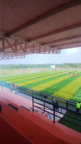 The camera pans across a soccer field with players running around, training or playing a game. The field is striped with alternating light and dark green turf. Spectators can be seen in the stands, some walking around and others lying down. A few people in bright yellow vests are standing near the railing. The stands are red with blue metal railings. The stands have a white wooden roof structure overhead. The background shows trees, buildings, and mountains under a cloudy sky.