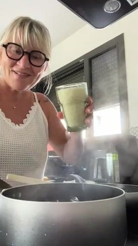 A middle-aged woman with blonde hair and glasses is in a kitchen. She is smiling and holding a glass of uncooked rice, pouring it into a pot of steaming water. She gives a thumbs-up to the camera. The kitchen appears to be modern, with stainless steel appliances and blinds on a window in the background. There is steam rising from the pot, indicating that the water is boiling.
