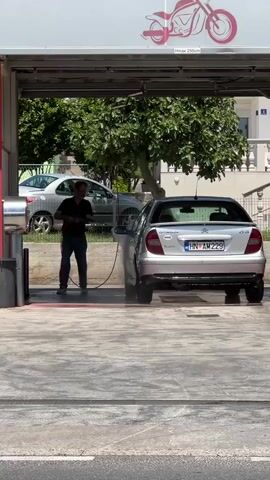 A man is pressure washing a silver car at an outdoor car wash. He is wearing a black t-shirt and blue jeans, and he is holding a pressure washer wand with a red hose attached. The car is parked under a covered bay, and there are trees and other cars in the background. The man is aiming the pressure washer at the rear passenger side of the car. The ground is wet and stained with red soap.