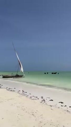The video shows a white sand beach with calm, shallow, turquoise water, under a partly cloudy sky. Several people are walking along the beach in the distance. Two small, traditional wooden sailboats, each with a single triangular sail, are beached near the water's edge. A person is standing near one of the boats. There is some seaweed debris scattered along the shoreline.