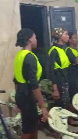 The video shows a group of women standing inside a room with blue walls and a clock on the wall. The women are wearing black tops with bright green vests and black skirts. The skirts seem to be embellished with shiny decorative elements. They are gathered around light-colored chairs with a distinctive design. Some of the women are singing, while others are standing around and observing. There are additional people in the background who are partially visible. The overall setting appears to be indoors, and the lighting is slightly dim.