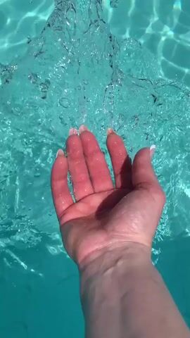 A hand is submerged in turquoise water, its fingers cupped. Water flows over and around the hand, creating ripples and splashes. The scene is shot from a close-up perspective, focusing on the interaction between the hand and the water. The lighting suggests a bright, sunny environment.