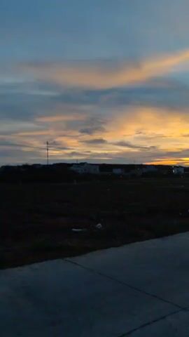 The video shows a sunset over a field with a few buildings in the distance. The sky is a mix of blue, orange, and gray clouds. In the foreground is a concrete road. A motorcycle with two people on it drives past the camera. A person is holding a smartphone, presumably to take a picture of the scene.
