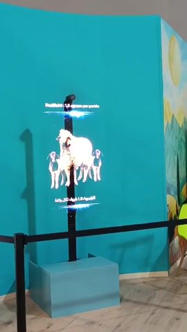 A holographic display system showcases animated images of sheep and lambs against a vibrant teal wall in Al Machouar – Stinia, Morocco. The holographic figures appear to float, transitioning from a mother sheep with two lambs, dissolving into particles, and then reforming as two sheep before dissolving again. Text in both French and Arabic, partially visible above and below the hologram, states "Prolificité : 1.6 agneaux par portée" (Prolificacy: 1.6 lambs per litter). The display unit consists of a black pole emerging from a light blue rectangular base. A black stanchion with a rope forms a barrier in front of the display. To the right, a colorful mural depicting mountains and a sun is visible on the wall.