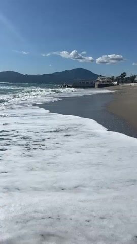 The video shows a sunny day at the beach. Waves with white foam roll onto the dark sand. In the background, there are mountains and a building, possibly a restaurant or hotel. The sky is blue with a few white clouds.