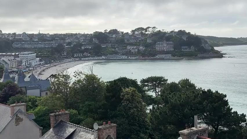 A wide shot overlooks a coastal town with a bay and a beach. The sky is overcast. In the foreground, rooftops and trees are visible. The middle ground features a curved sandy beach with several people scattered along it, and a line of buildings and trees bordering the beach. Beyond the beach, the bay is filled with calm water and several boats, including sailboats. The background shows a hilly coastline covered in houses and trees, stretching into the distance along the sea.