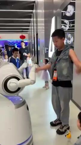 A young boy interacts with a white robot. The robot, with a round body and two arms, stands on a white floor in a well-lit indoor space. The boy, wearing a sleeveless jean jacket and gray pants, gestures towards the robot, and the robot raises its arms in response. Other children and adults are visible in the background, suggesting a public or commercial setting. The environment includes elements of modern design with sleek surfaces and decorative lighting.
