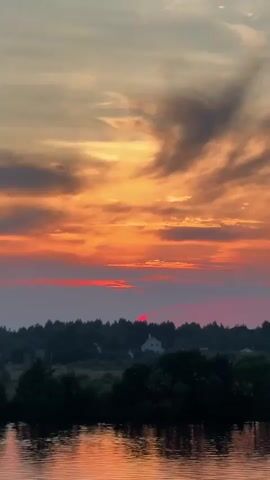 The video shows a sunset over a body of water with trees in the foreground. The sky is filled with vibrant orange and red clouds, and the sun is setting behind a line of trees. A white house is visible in the distance, nestled among the trees. The water reflects the colors of the sky, creating a beautiful and serene scene.
