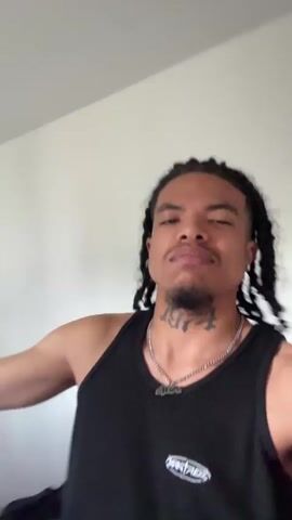 A medium shot shows a person with dark hair in braids, wearing a black tank top and a silver chain with a pendant, standing in what appears to be a bedroom. They are looking directly at the camera and speaking, making gestures with their hands. A bed with a grey headboard and some items on it is visible in the background, along with a white wall. The lighting in the room appears natural.