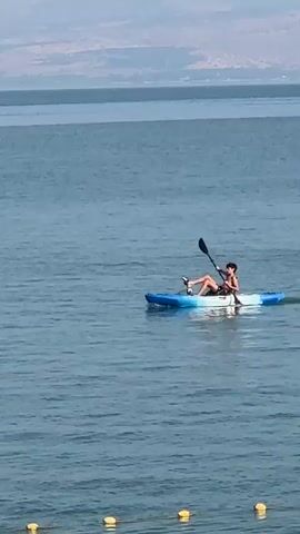 A person is kayaking on a large body of water. The person is wearing a dark swimsuit and is using a black paddle to propel the kayak. The kayak is blue and white. The water is calm and blue. In the background, there is a distant shoreline with hills. The sky is clear and hazy.