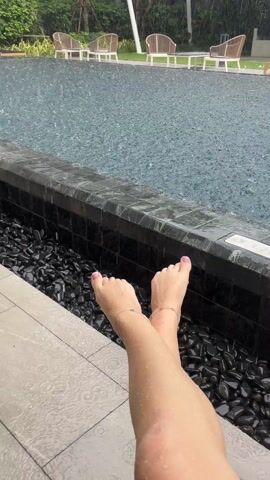 The video shows a pair of fair-skinned legs, each adorned with a delicate silver anklet, extended towards a swimming pool on a rainy day. The toes are painted a light pink.  The pool is surrounded by dark, smooth stones, and a tiled edge. Rain falls heavily on the surface of the pool, creating numerous visible droplets. In the background, there are several chairs and tables arranged on a grassy area, some are sheltered by a white awning. Trees and buildings are visible in the distance, suggesting an urban or suburban setting. The overall impression is one of relaxation despite the inclement weather.