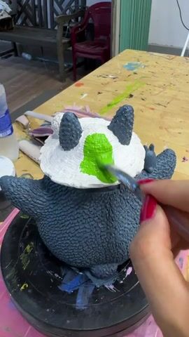A person's hand with red fingernails is painting a green area on the white face of a dark grey ceramic figure that resembles a panda or a similar creature. The figure has black ears and is positioned on a black turntable. The background shows a wooden surface with paint splatters and various bottles and brushes. The scene appears to be an art or craft studio.