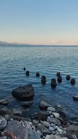 The video displays a serene seascape. The foreground is occupied by a rocky shoreline composed of various sizes of stones, ranging from small pebbles to larger boulders. Some of these rocks are partially submerged in the clear, blue water. In the water, there are several dark, weathered pilings or posts protruding from the surface, arranged in a somewhat scattered pattern. These structures suggest the remnants of an old pier or jetty. Beyond the pilings, the vast expanse of the ocean stretches towards the horizon. The water is calm with gentle ripples. In the distance, a faint, hazy outline of mountains or hills can be seen against the backdrop of a clear, pale blue sky. The lighting suggests it is either early morning or late afternoon, with soft, diffused light. There are no people or animals visible in the frame. The overall atmosphere is peaceful and tranquil.